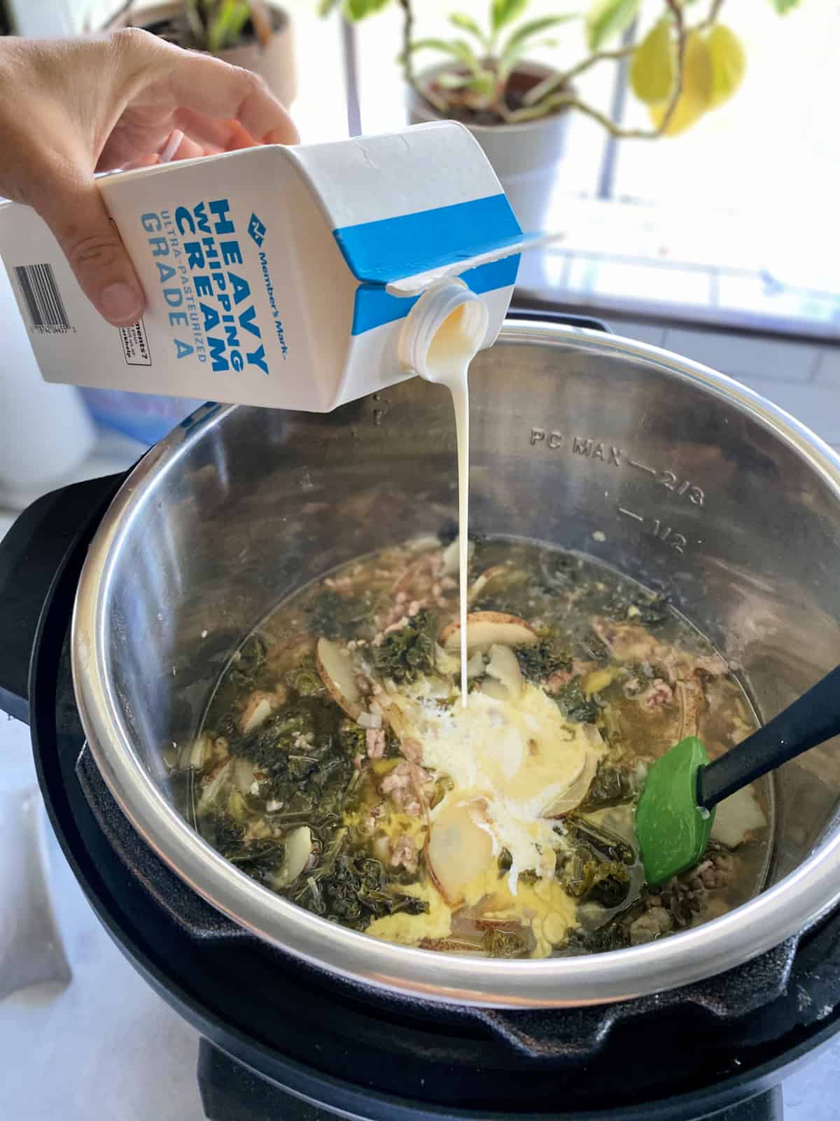 Hand pouring heavy cream into an Instant Pot full of soup with a green spatula.