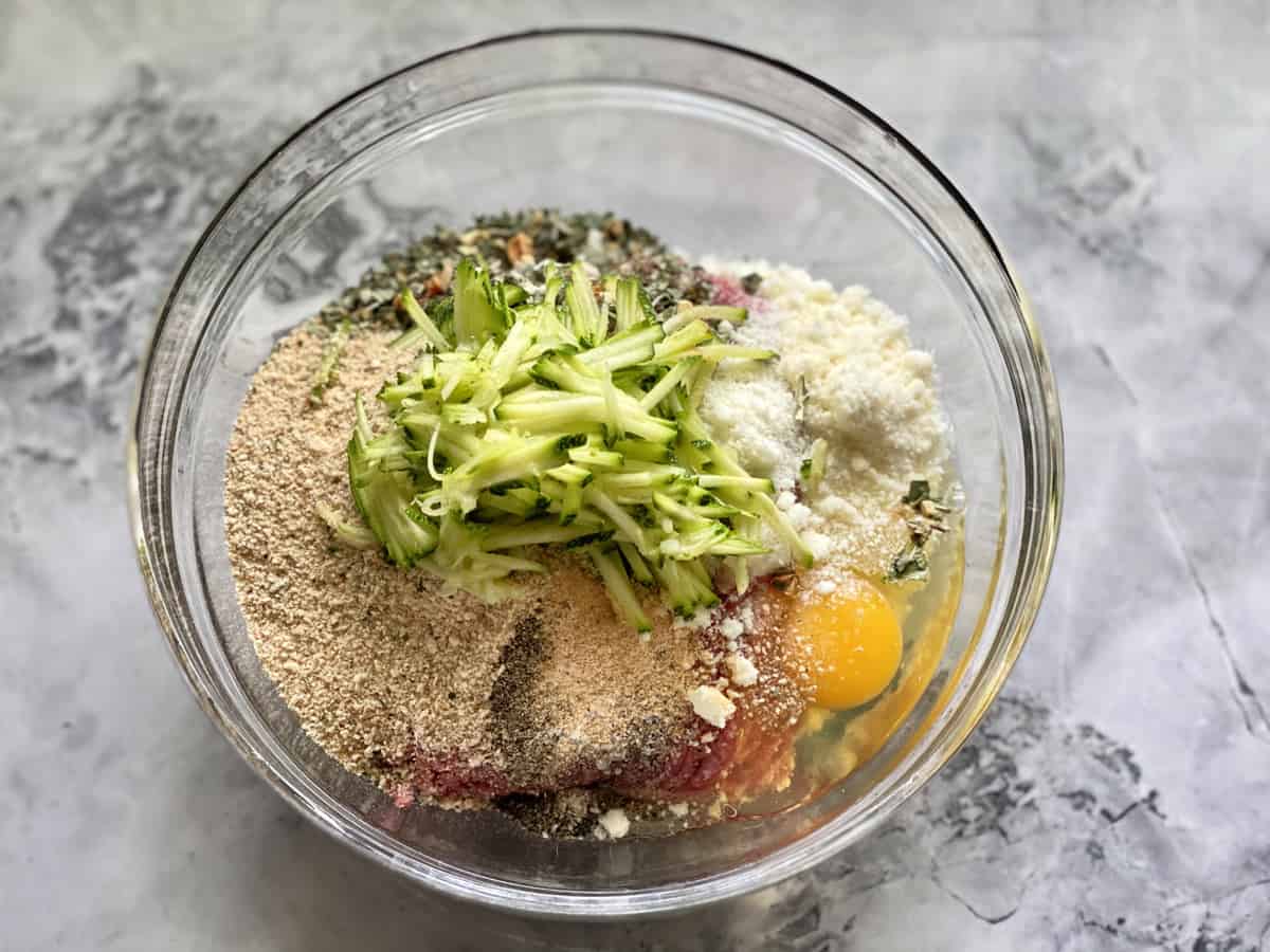 Glass bowl on countertop filled with ground beef, breadcrumbs, cheese, egg, and zucchini.