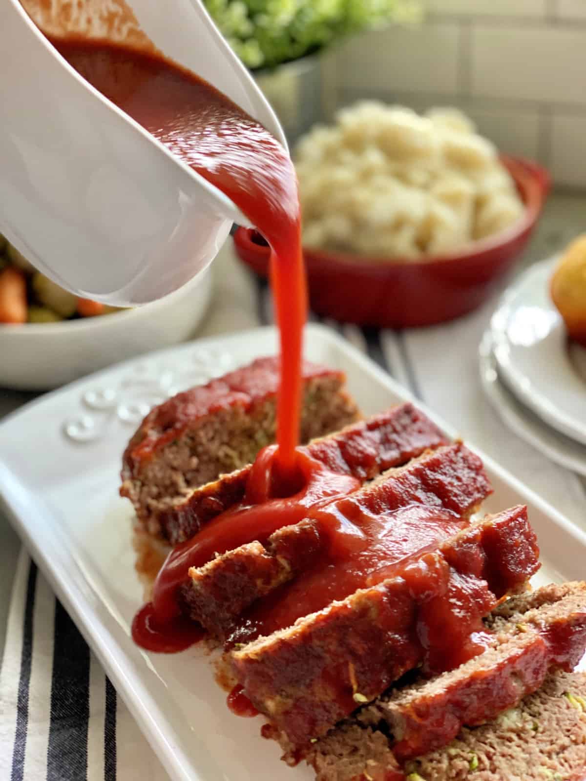 White gravy boat pouring red sauce on top of sliced meatloaf.