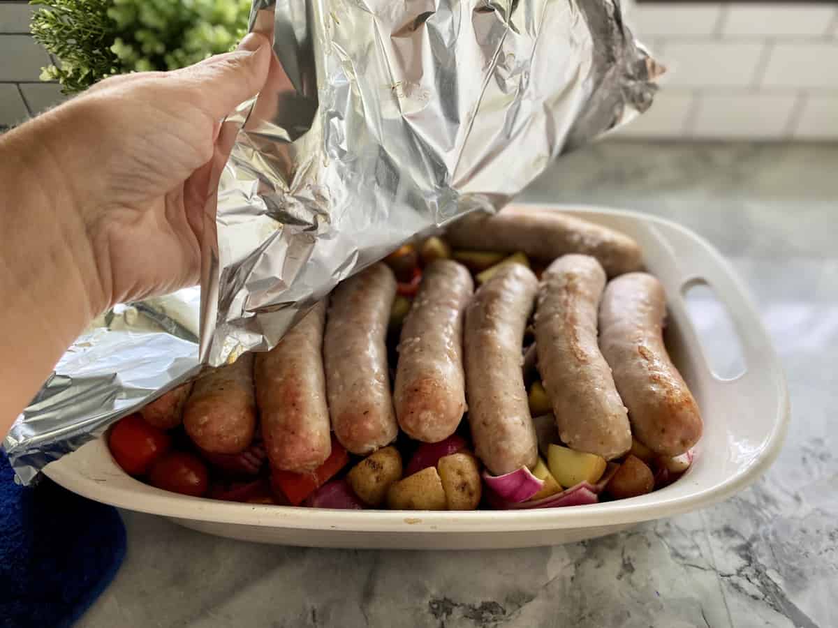 Hand pulling aluminum foil off pan loaded with Italian Sausage, potatoes, and vegetables. 
