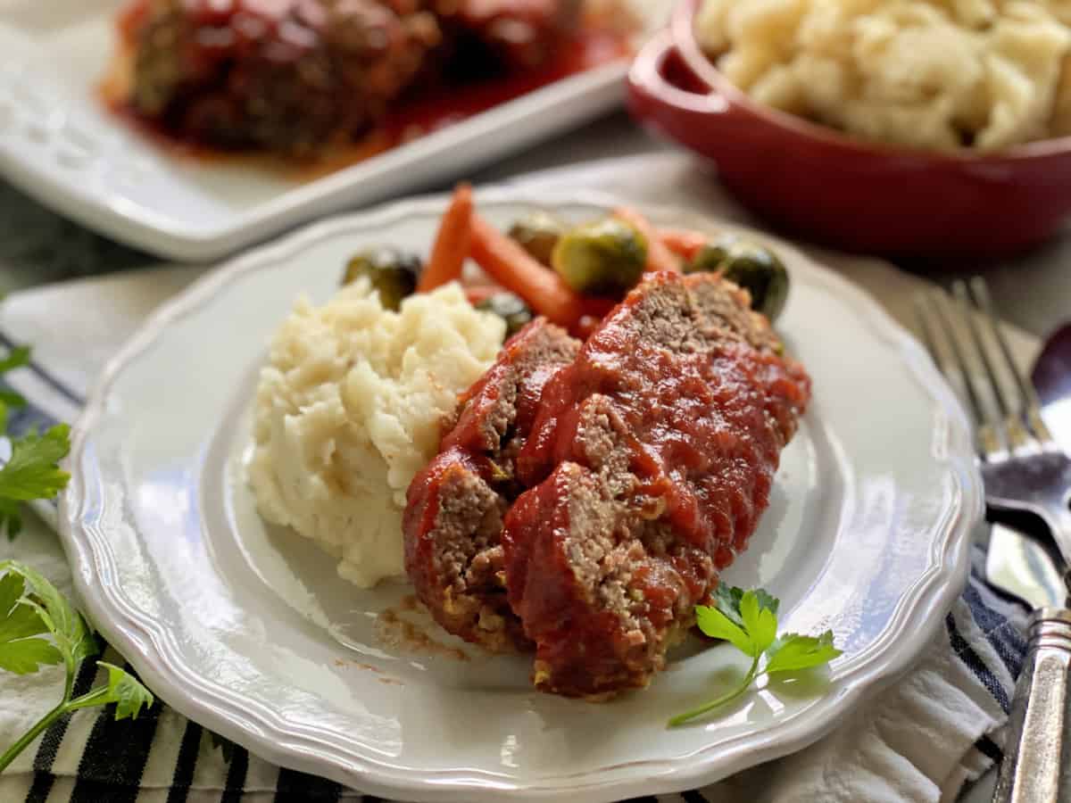 White plate with two slices of meatloaf with tomato sauce and mashed potatoes.