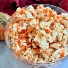 Glass bowl filled with orange popcorn with fall leaves and pumpkins around it.