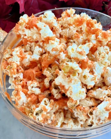 Glass bowl filled with orange popcorn with fall leaves and pumpkins around it.