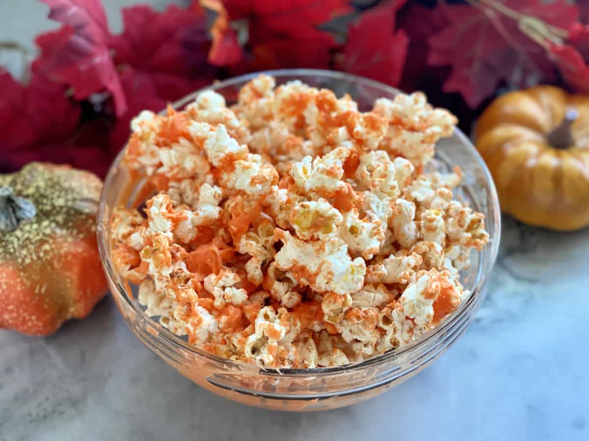 Glass bowl filled with orange popcorn with fall leaves in background.