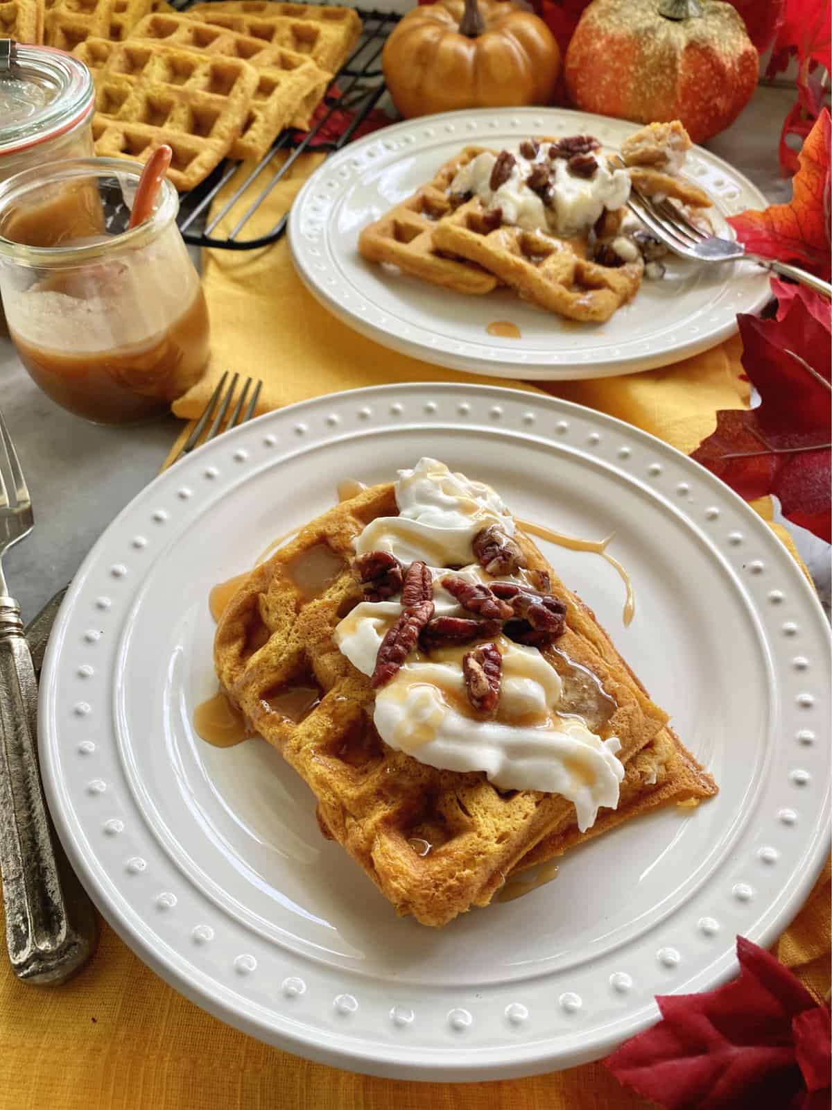 Two white plates with waffles topped with whipped cream, caramel sauce, and pecans.