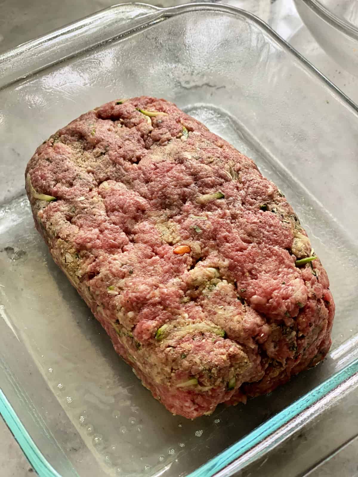 Ground beef shaped into a loaf sitting in a greased glass pan on a marble counter.