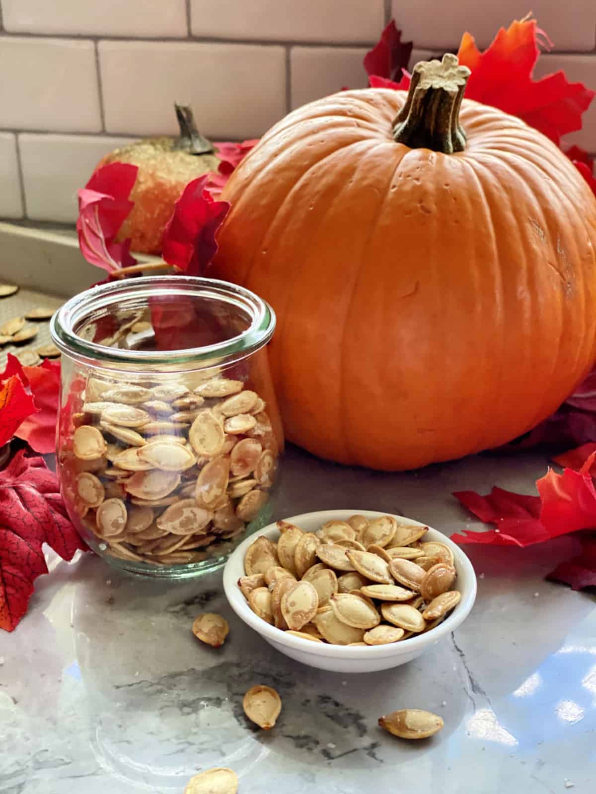 Roasted pumpkin seeds in a little white bowl, glass jar and a pumpkin in background.