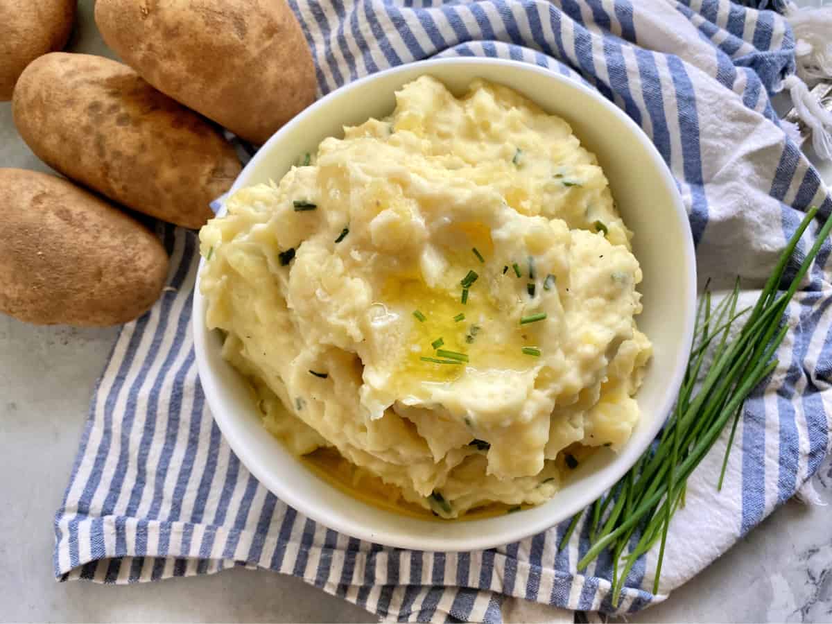 Overhead photo of a bowl of mashed potatoes with chives and baking potatoes.