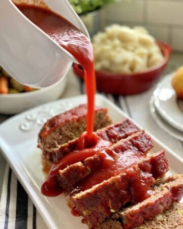 White gravy boat pouring red sauce on top of sliced meatloaf.