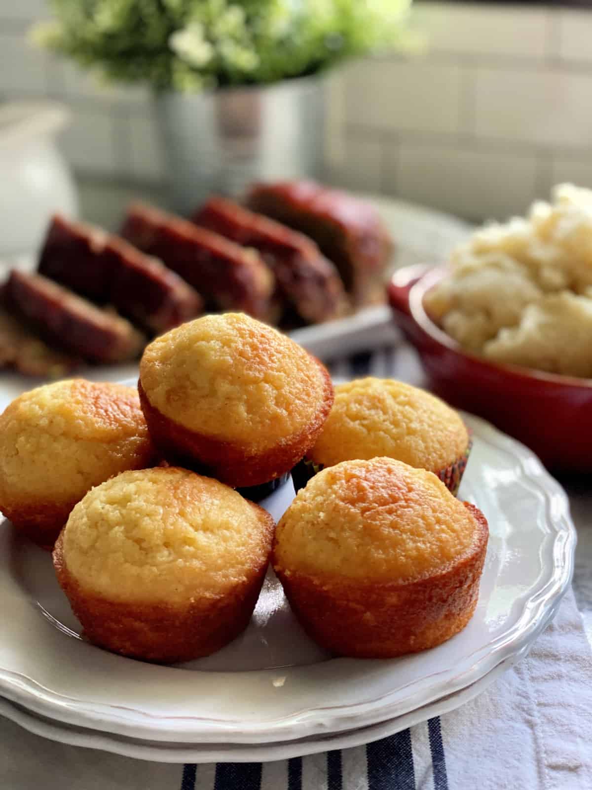 Six cornbread muffins stacked and sitting on two white plates with meatloaf in the background.