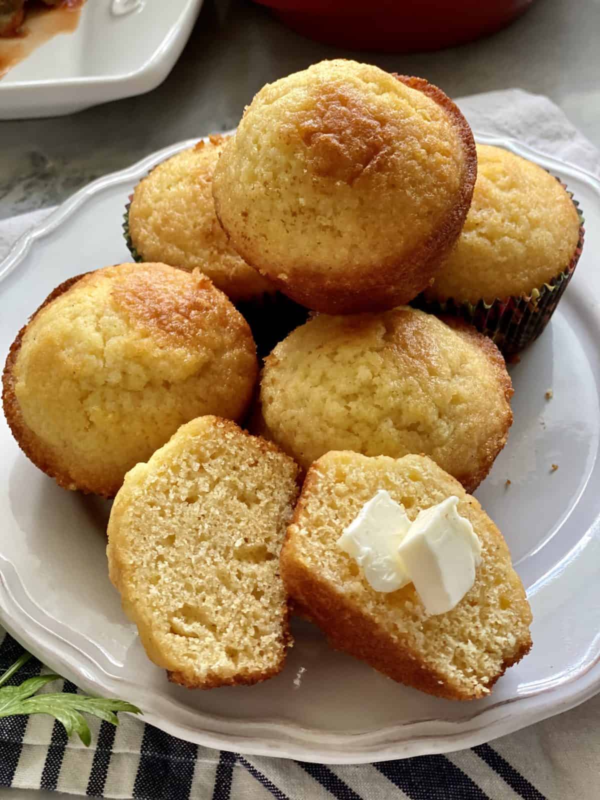Cornbread muffins stacked on a white plate with one muffin split in half with butter on it.