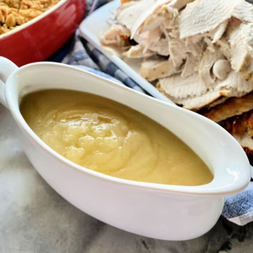 White gravy boat with yellow gravy inside and turkey in the background.
