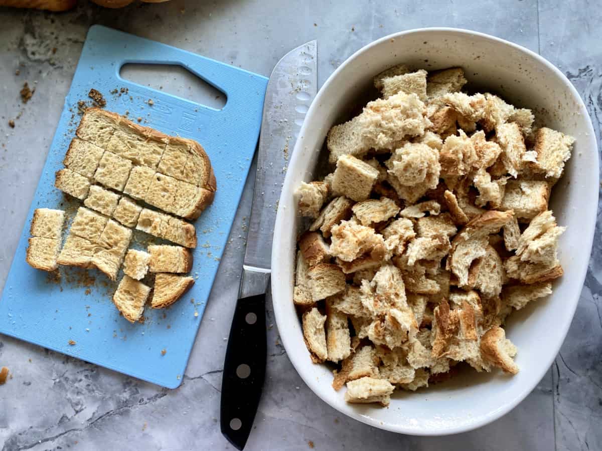 Top view of a casserole dish filled with cubed toasted bread and a cutting board next to it with cubed bread.