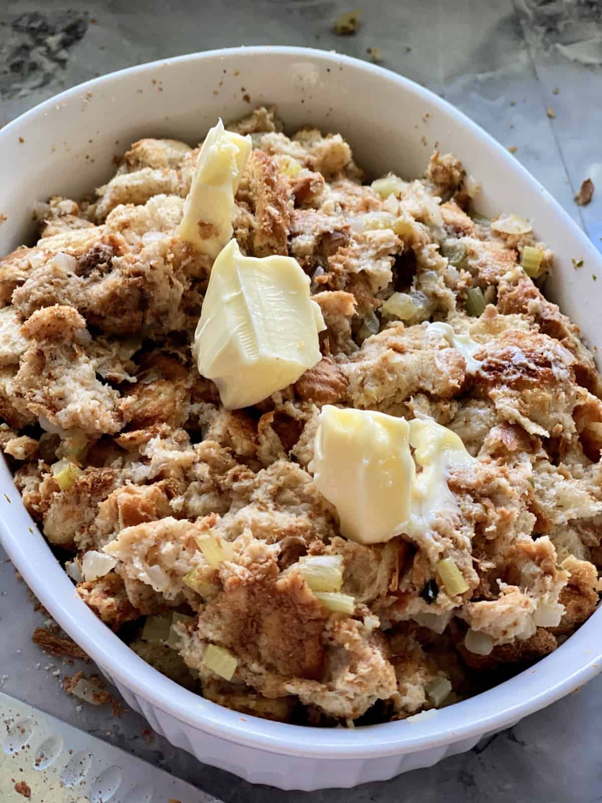 White casserole dish with bread stuffing and chunks of unmelted soft butter on top.
