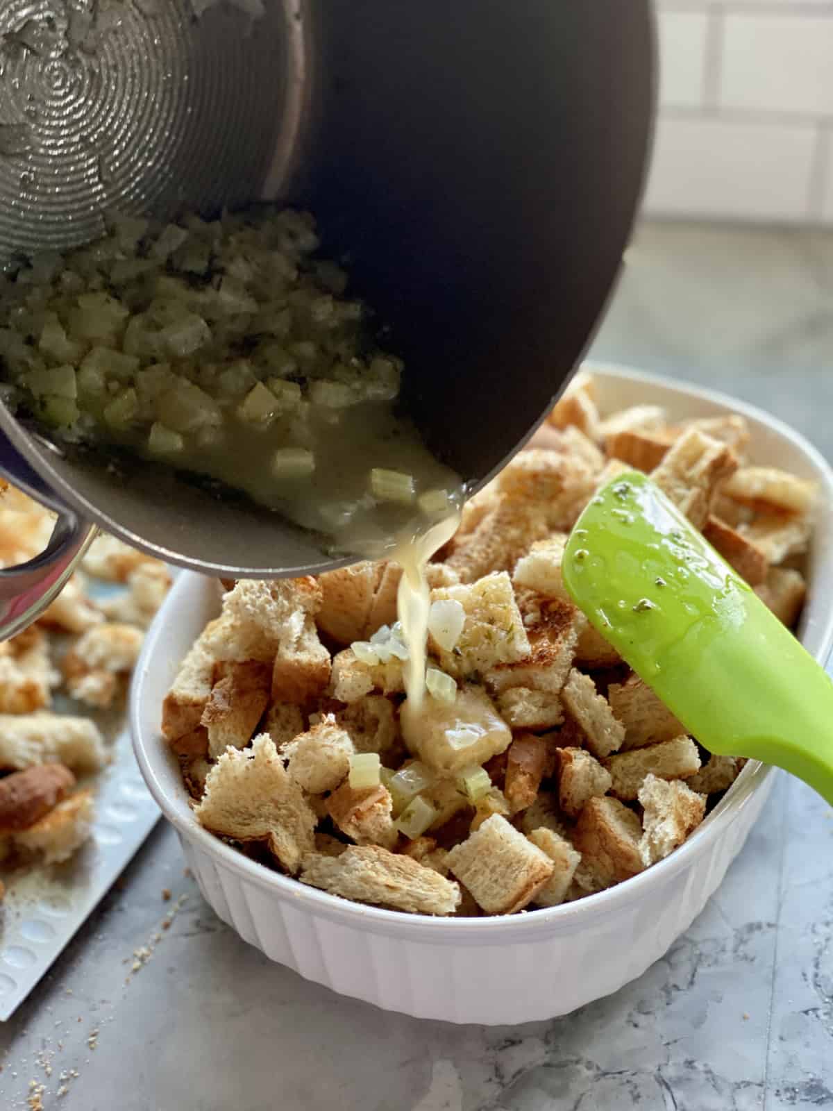 Brown pot pouring broth with celery into a white casserole dish filled with cubed bread.