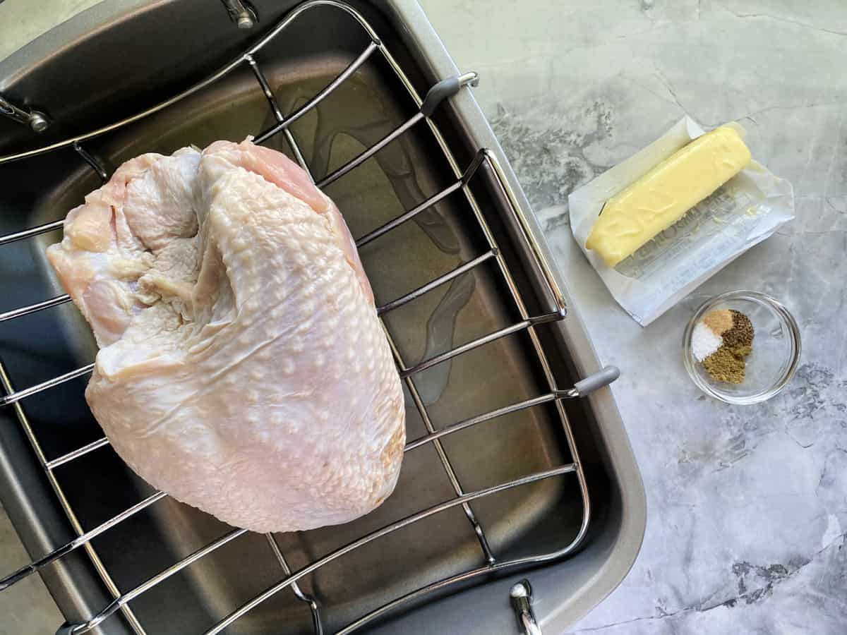 Raw chicken breast on a roasting rack with a stick of butter and herbs.