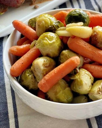 White bowl filled with roasted baby carrots, garlic cloves, and brussels sprouts.