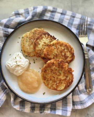 Top view of four mashed potato cakes with sour cream and applesauce on a checkerred cloth.