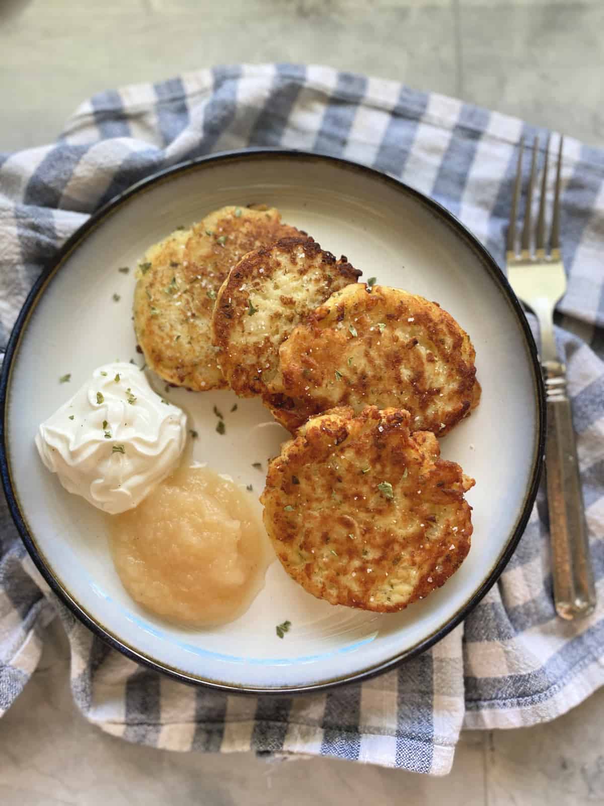 Top view of four mashed potato cakes with sour cream and applesauce on a checkerred cloth.