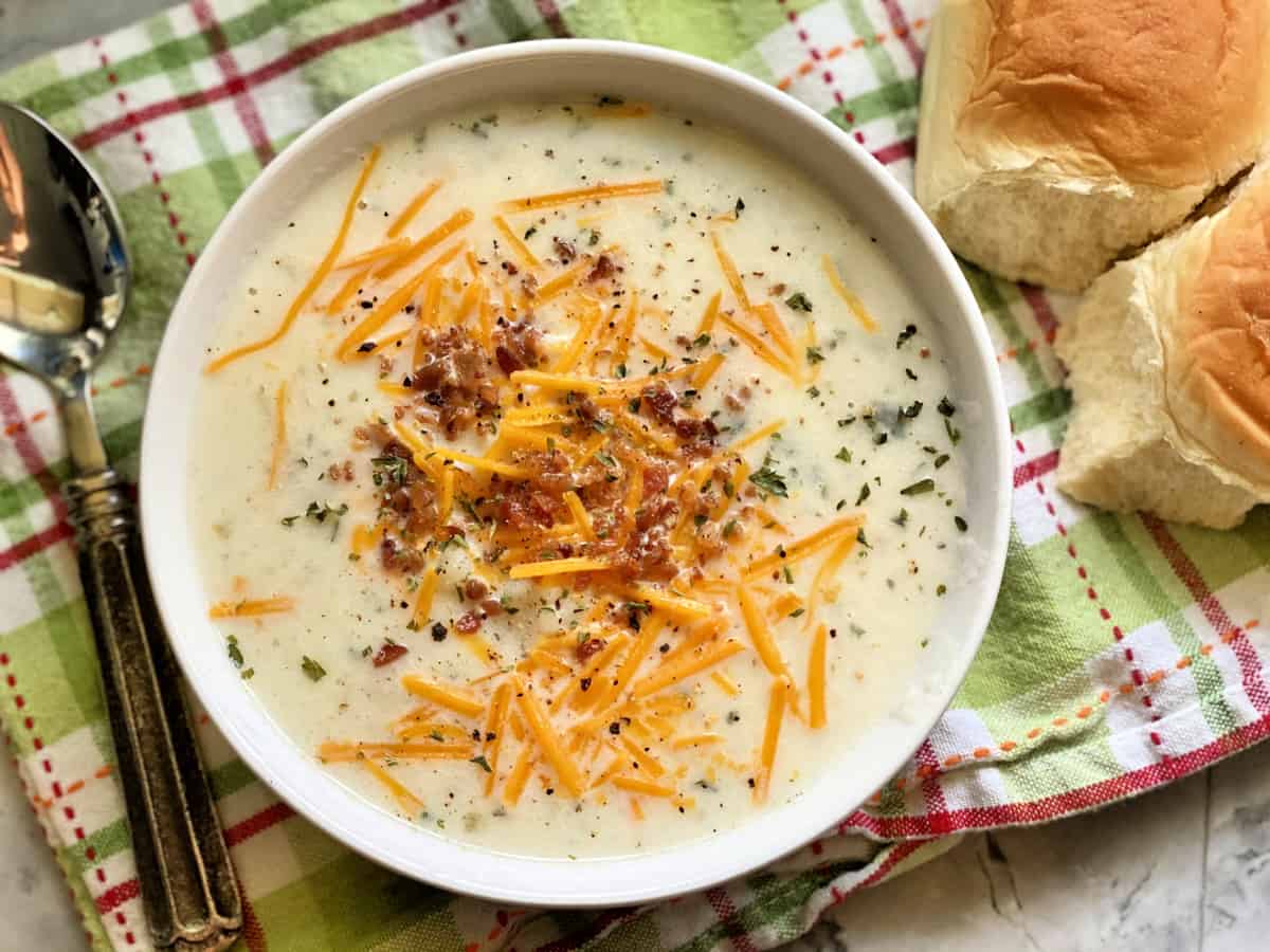 Top view of a bowl of potato soup with cheese, bacon, and herbs with two rolls next to the bowl.