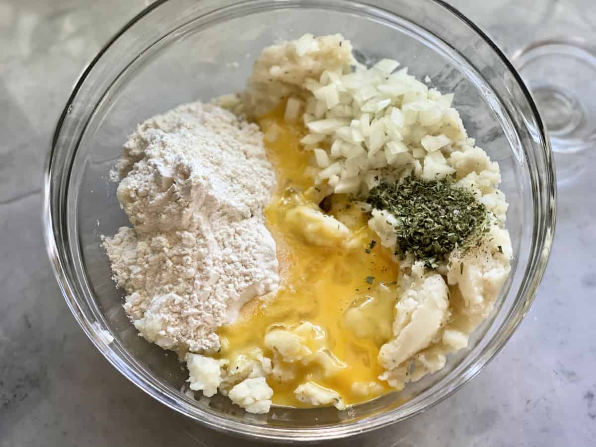 Glass bowl filled with flour, egg, diced onion, seasonings, and mashed potatoes.