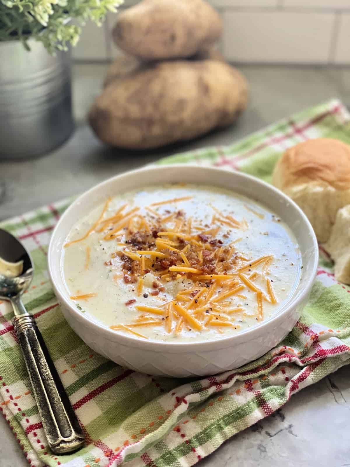 A single bowl of potato soup topped with bacon and cheese with potatoes in background.
