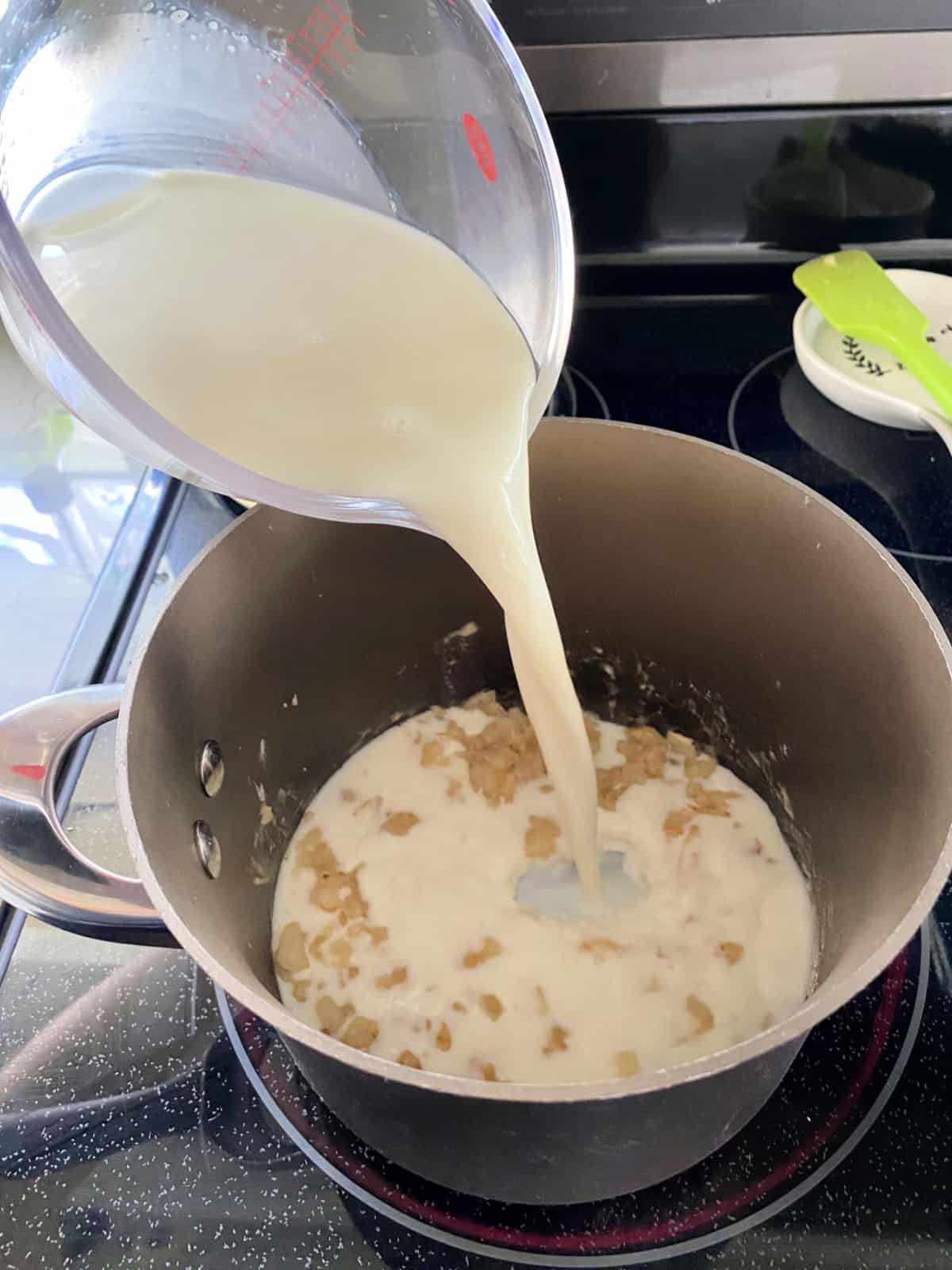 Measuring cup filled with milk being poured into a saucepan with onions.