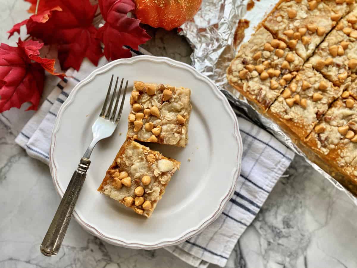 Two pumpkin pie bars on a white plate with a fork sitting on a dish towel.