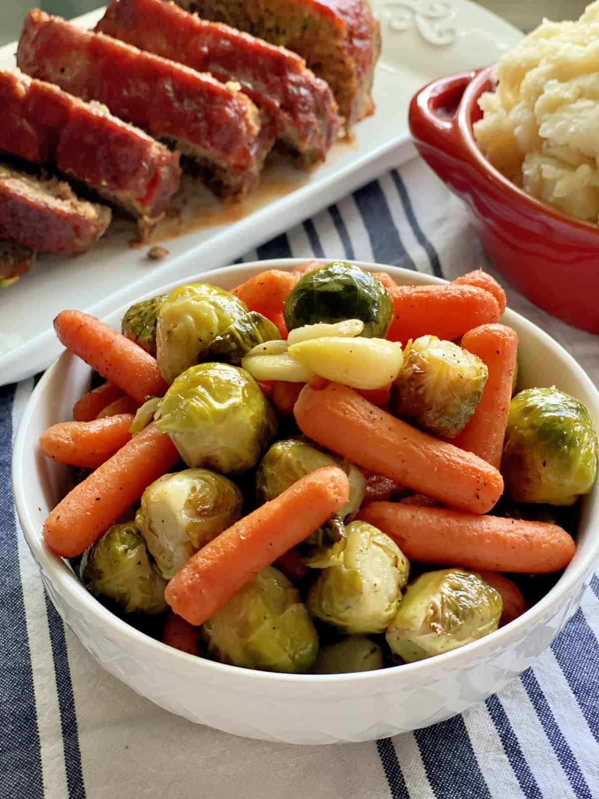 White bowl filled with roasted baby carrots, garlic cloves, and brussels sprouts.