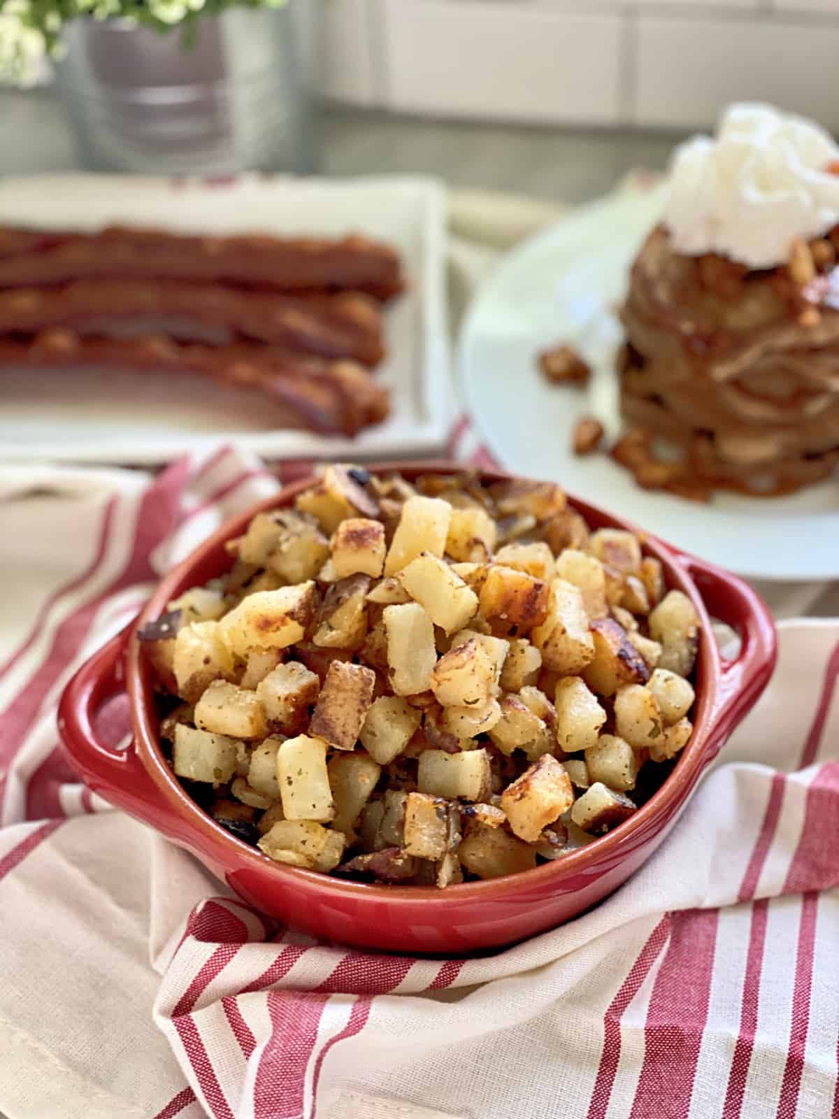 Bowl of diced cooked potatoes on a red and white striped cloth with pancakes in the background.