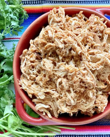 Top view of a red bowl filled with shredded chicken with fresh cilantro on the side.