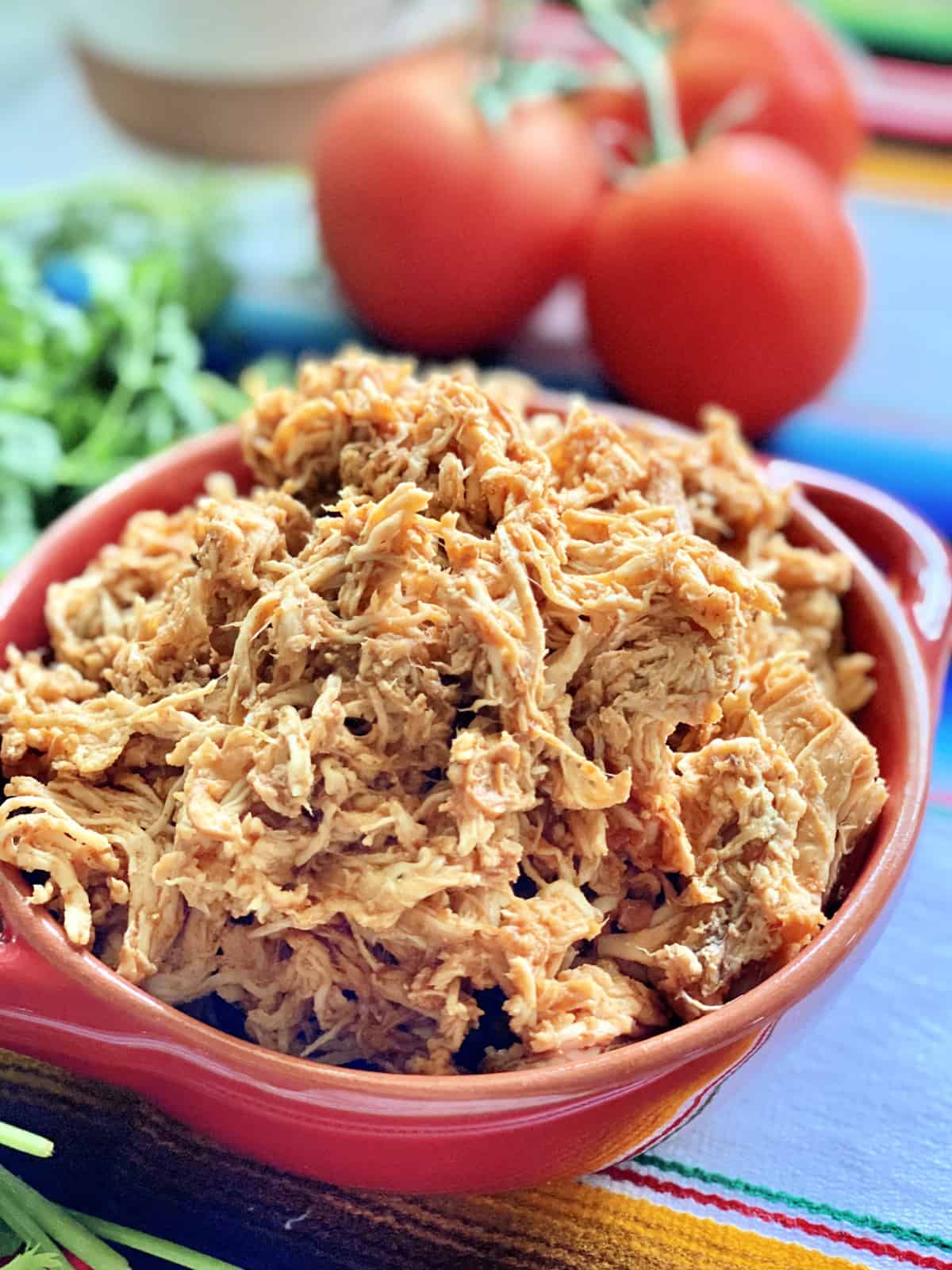 Red bowl filled with shredded chicken with tomatoes on the vine in the background.