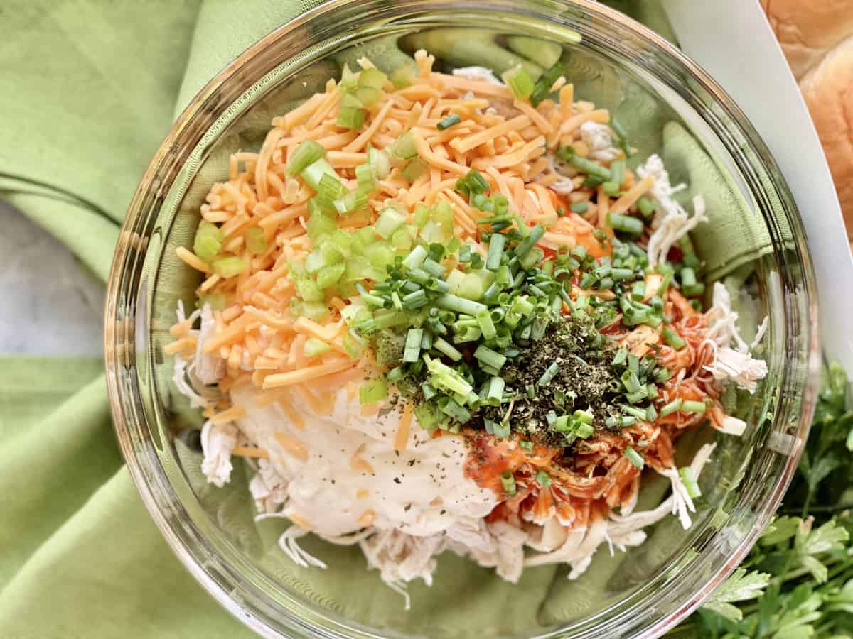 Top view of a bowl filled with shredded chicken, mayonnaise, cheese, buffalo sauce, spices, and greemn onions.