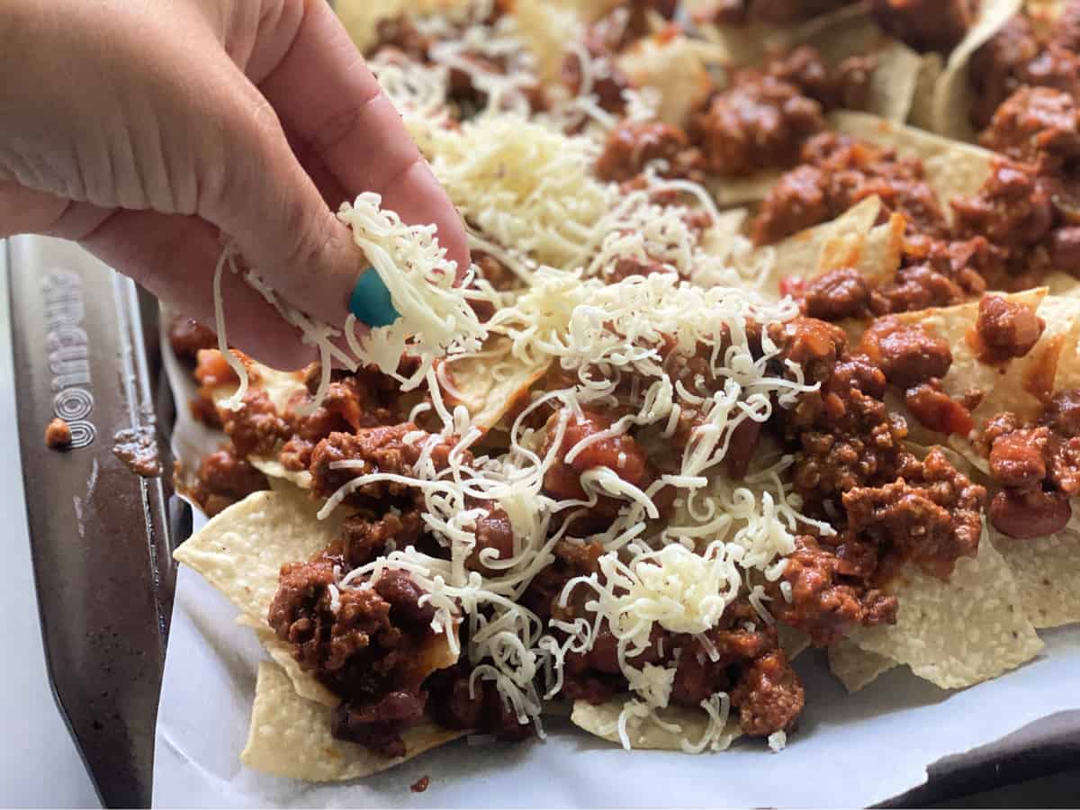 Hand sprinkling cheese over chili on nachos in sheet pan.