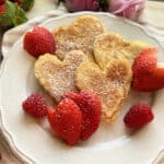 Four heart shaped pancakes on a white plate dusted with powder sugar and heart shapped strawberries.