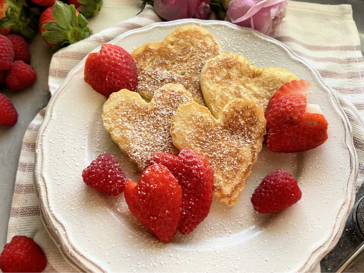 Four heart shaped pancakes on a white plate dusted with powder sugar and heart shapped strawberries.