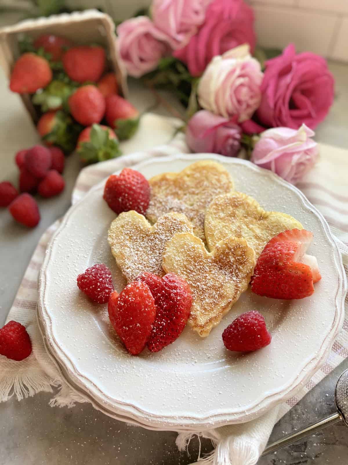 Four heart shaped pancakes on a white plate with heart shaped strawberries.