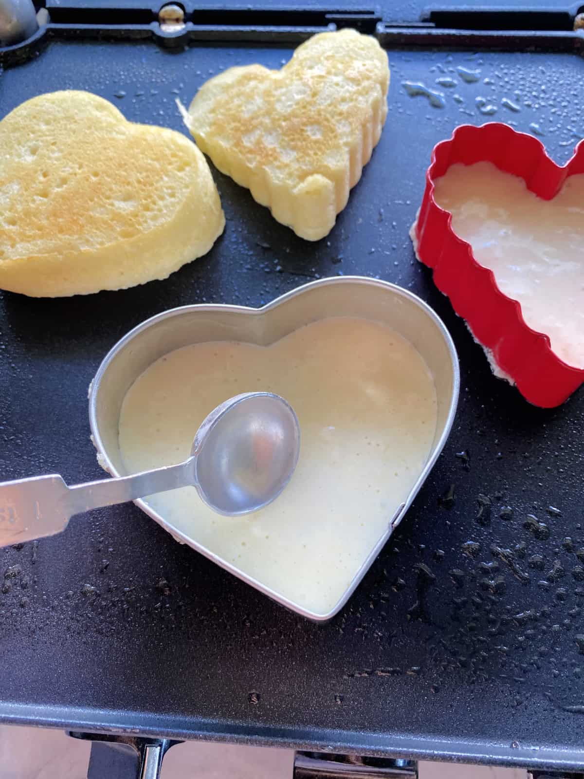 Measuring spoon moving batter to the sides of a heart shapped cookie cutter.