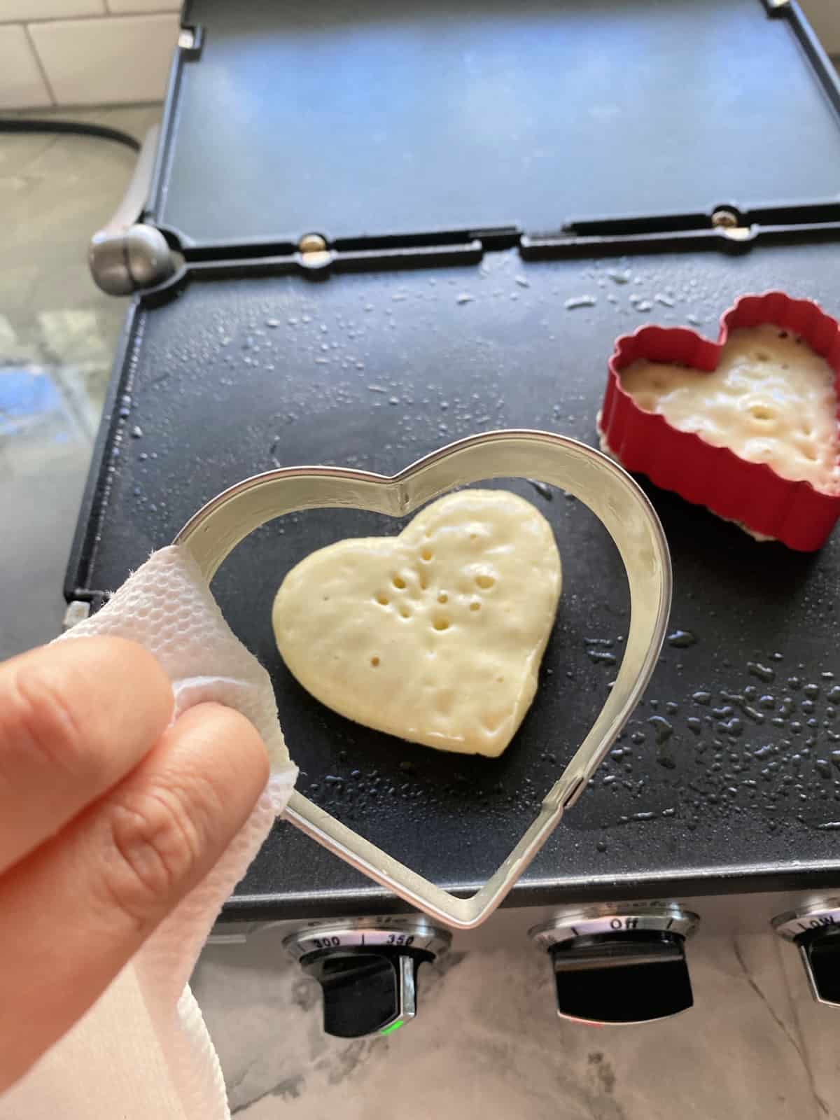 Hand holding a paper towel removing the heart shaped cookie cutter from the griddle.