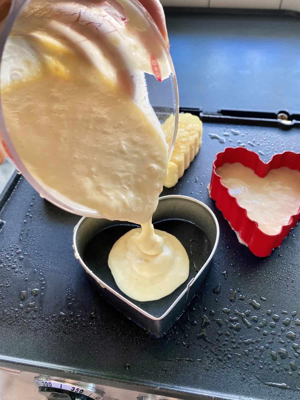 Measuring cup pouring pancake batter into a heart shaped cookie mold on a griddle.