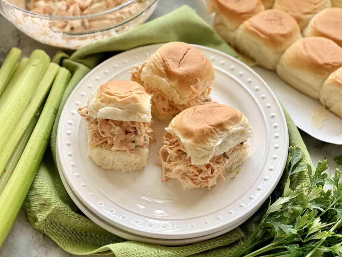 Three chicken salad sandwiches on a white plate with rolls and celery in the background.