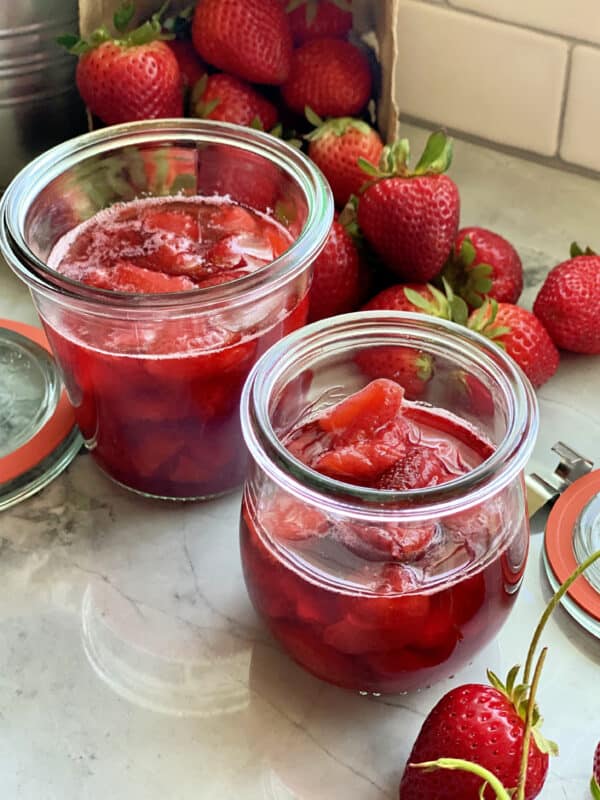 Two glass jars with chunky strawberry sauce and stemmed strawberries in the background.