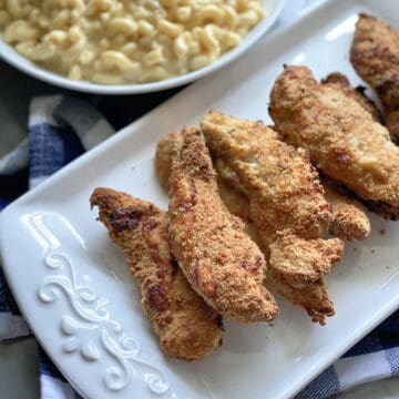 Top view of baked chicken strips on a white platter with a bowl of mac and cheese in background.