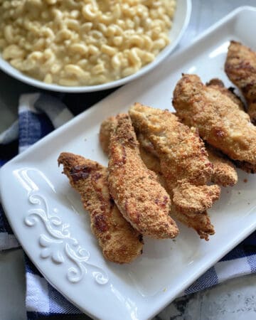 Top view of baked chicken strips on a white platter with a bowl of mac and cheese in background.