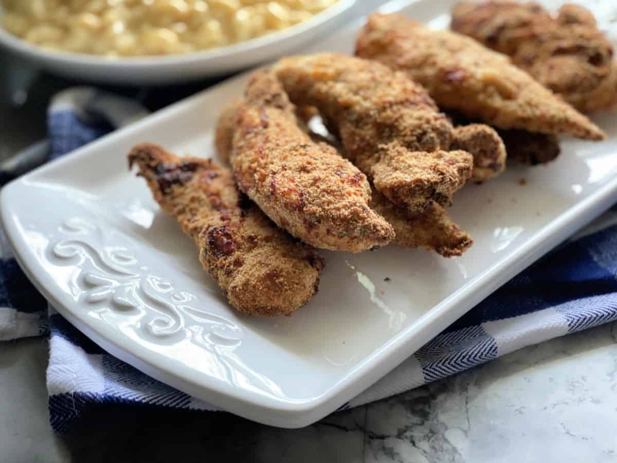 Chicken strips stacked on a white platter sitting on a blue and white checkered cloth.