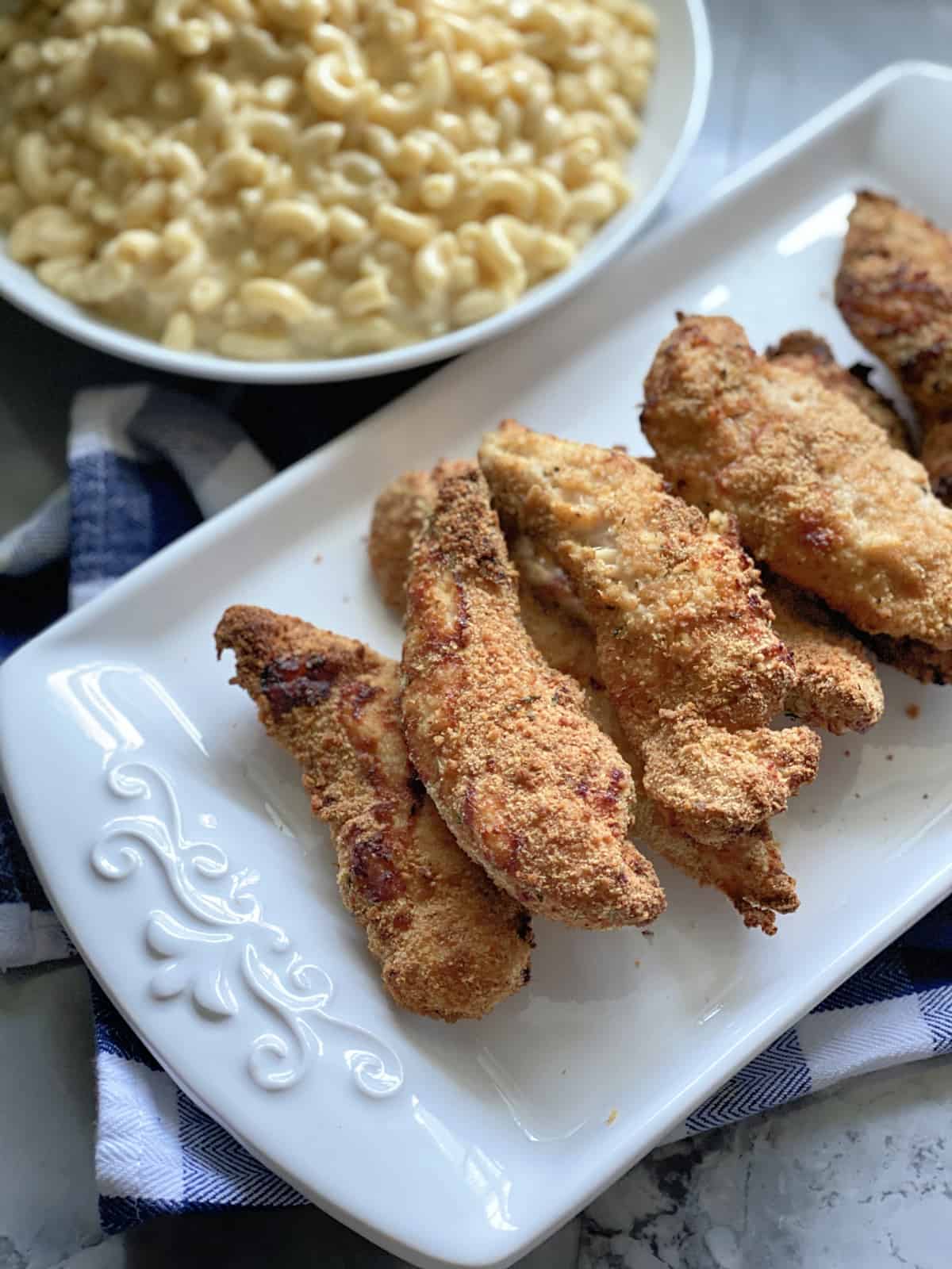 Top view of baked chicken strips on a white platter with a bowl of mac and cheese in background.