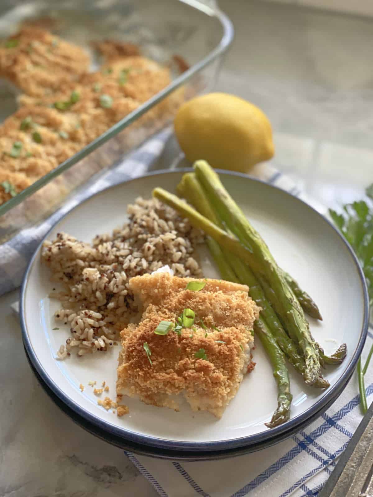 White plate with fish, rice, and asparagus on it with baked fish in background.