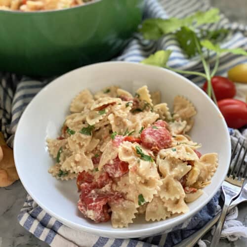 White bowl filled with bow tie pasta, tomatoes, and cream with a green pot in the background.