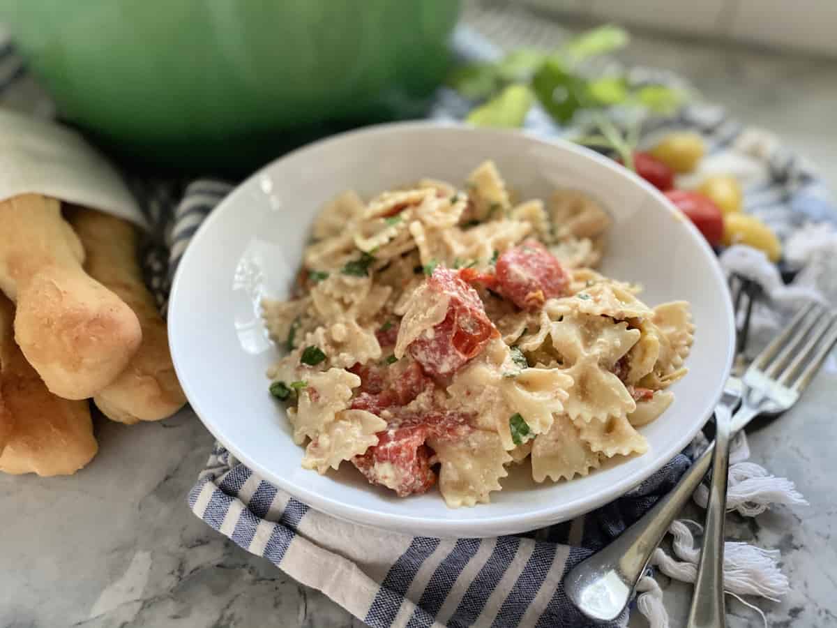 Bowl of bow tie pasta with smashed tomatoes and breadsticks on the side.