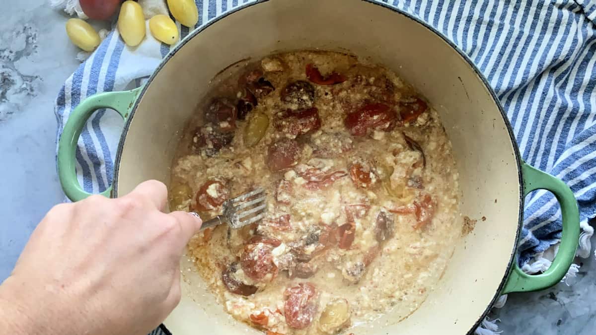 Female hand mixing baked feta and smashing blistered tomatoes with fork.